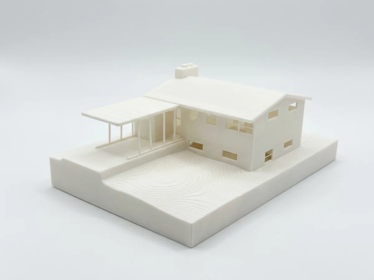 White 3D-printed model of a mid-century split-level house with a covered carport and chimney, sitting on a base with contour lines showing the yard